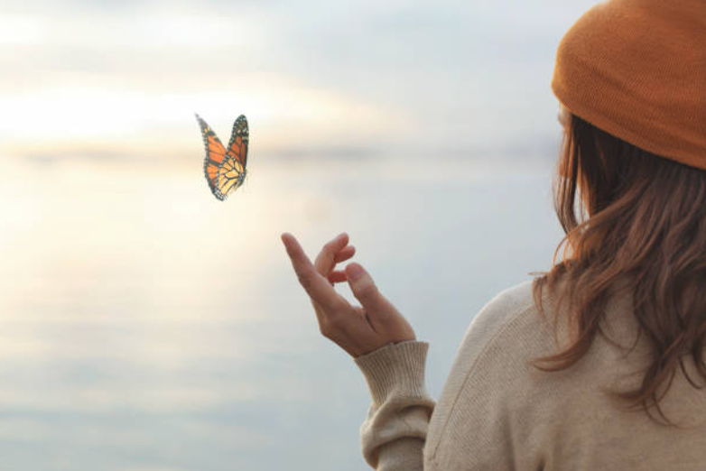 Chica de espaldas, frente al mar y con gorro naranja de punto. Tiene la mano izquierda extendida, y parece que una mariposa se va a posar en ella